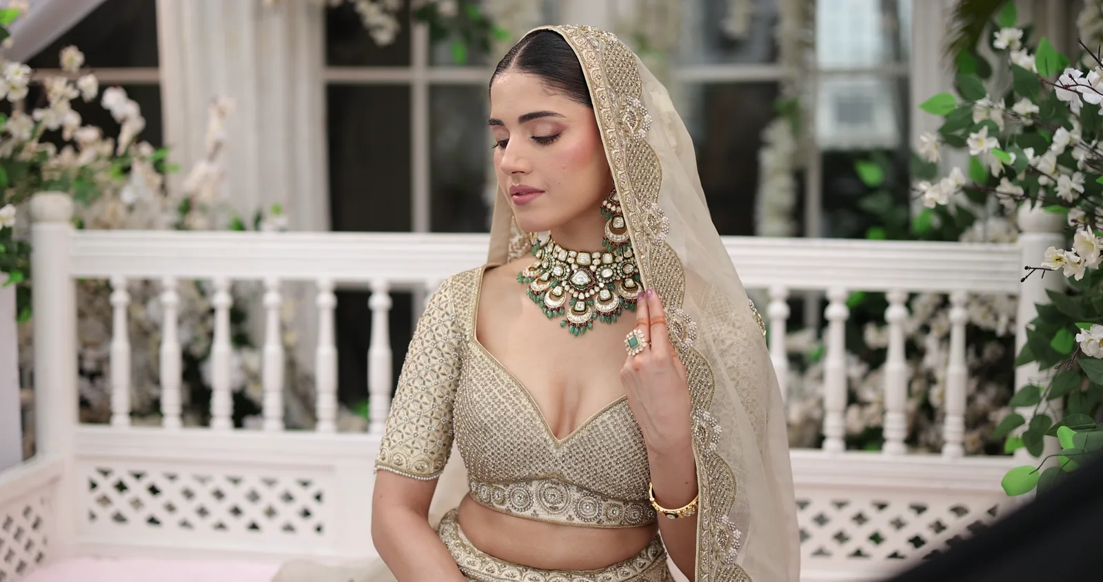 A bride in a polki-kundan choker with emerald drops, seated on a white pavilion bench under a sheer dupatta
